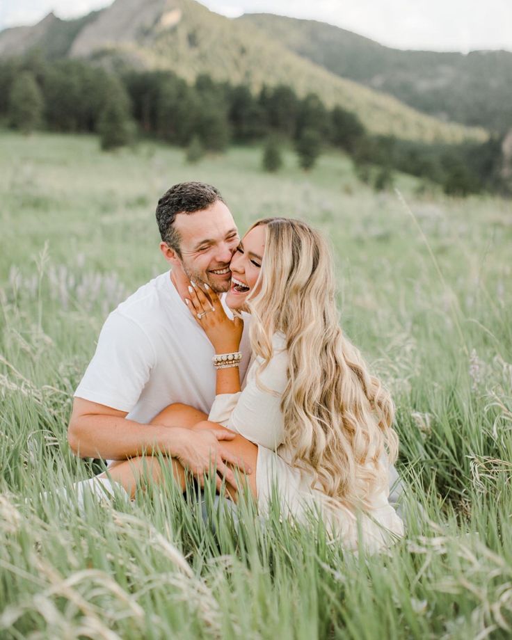 a man and woman are sitting in tall grass with mountains in the backgroud