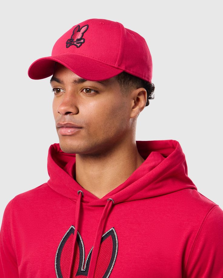 A person wearing the Psycho Bunny MENS WALTER BASEBALL CAP - B6A800D200 and a matching red hoodie with a bunny logo, looking to the side against a plain background. The durable and comfortable cap is crafted from 100% cotton, perfectly complementing the casual outfit. Casual Golf Visor Baseball Cap, Casual Baseball Cap For Golf, Casual Baseball Cap With Curved Brim For Golf, Casual Golf Baseball Cap With Curved Brim, Casual Baseball Cap With Curved Visor For Golf, Casual Curved Brim Baseball Cap For Golf, Casual Pink Snapback Hat For Sports Events, Cotton Trucker Hat For Baseball Sports, Cotton Trucker Hat For Baseball Season Sports