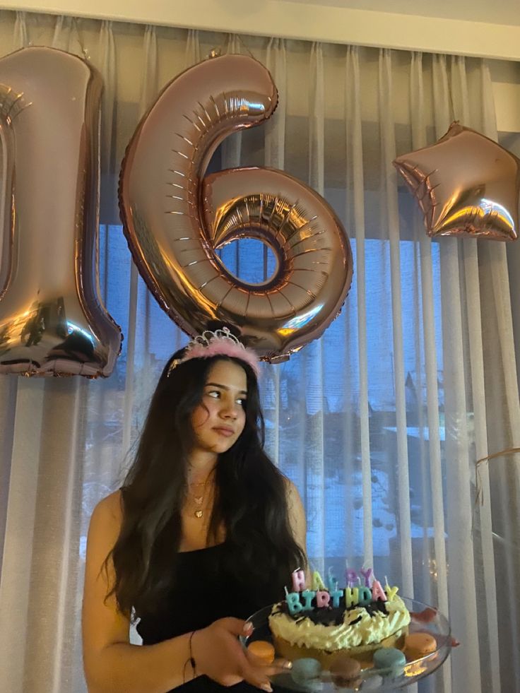 a woman holding a cake with candles on it in front of balloons that spell out the number six