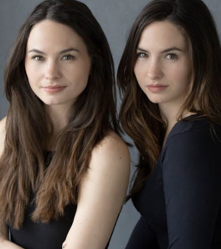 two beautiful women standing next to each other in front of a gray background with their arms crossed