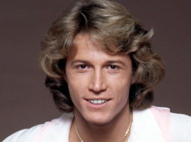 a man with blonde hair wearing a white shirt and gold necklace smiling at the camera