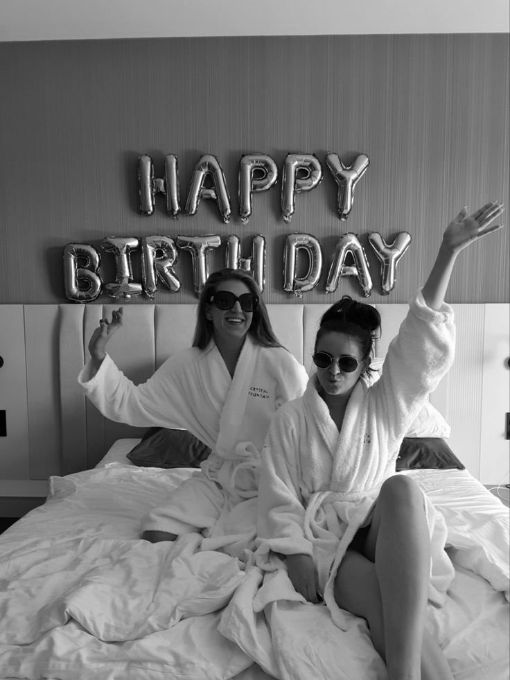 two women in robes sitting on a bed with the words happy birthday spelled above them