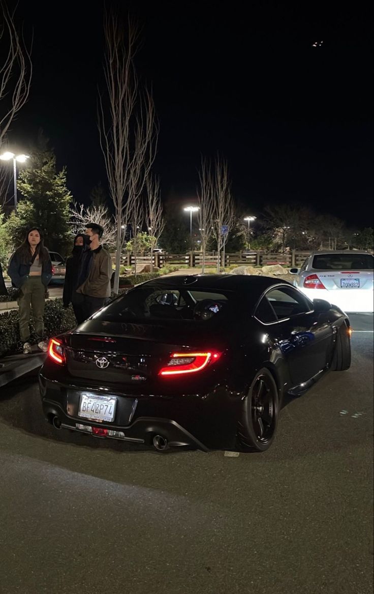 two people standing on the back of a black sports car in a parking lot at night