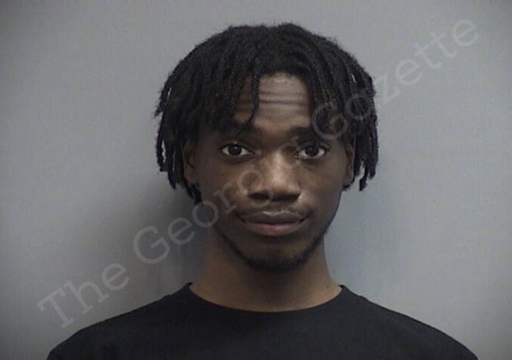 a man with dreadlocks is mugged in front of a gray wall and wearing a black t - shirt