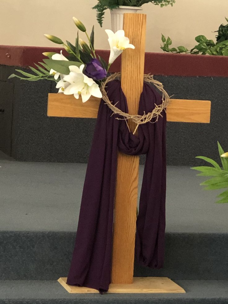 a wooden cross with flowers on it and a purple scarf draped around the cross, along with greenery