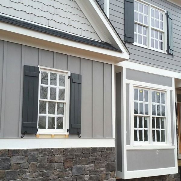 a gray house with two windows and shutters on each side, along with a stone wall