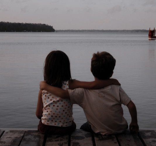 two children sitting on a dock looking out over the water at a boat in the distance