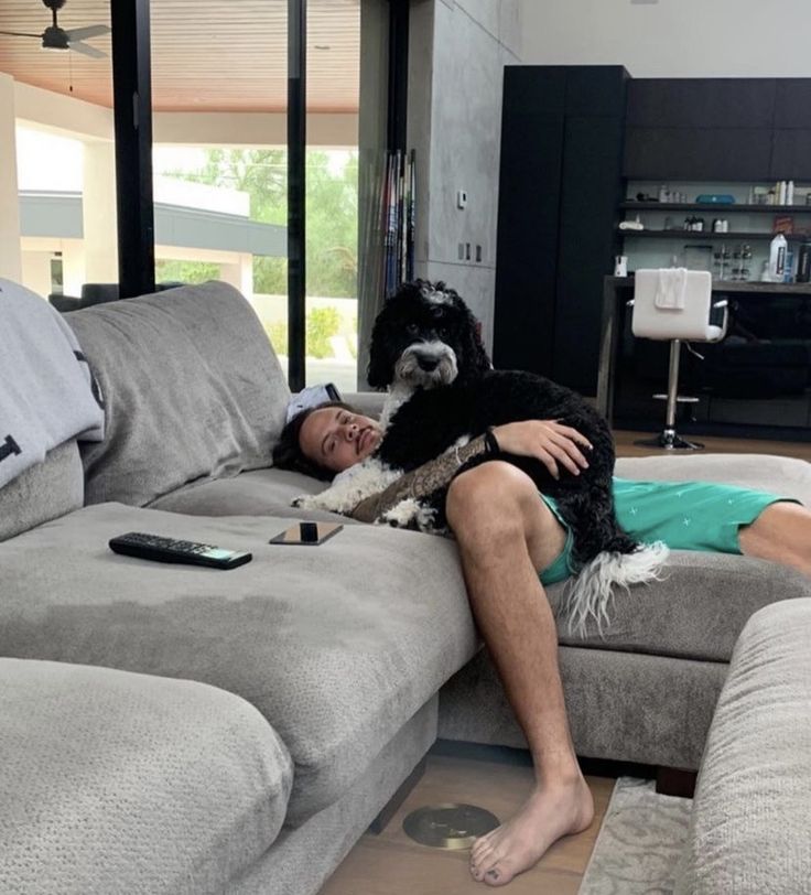 a man laying on top of a gray couch next to a black and white dog