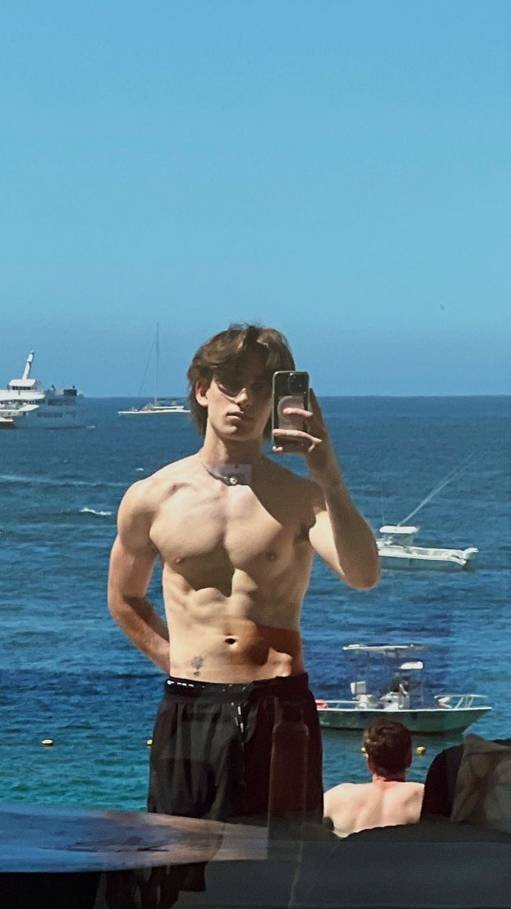 a shirtless man taking a selfie in front of the ocean with his cell phone