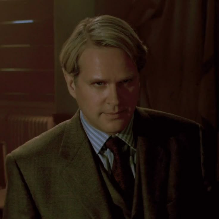 a man with blonde hair wearing a suit and tie in a dimly lit room, looking at the camera