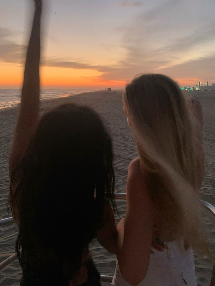 two women are sitting on the beach watching the sun go down with their arms in the air