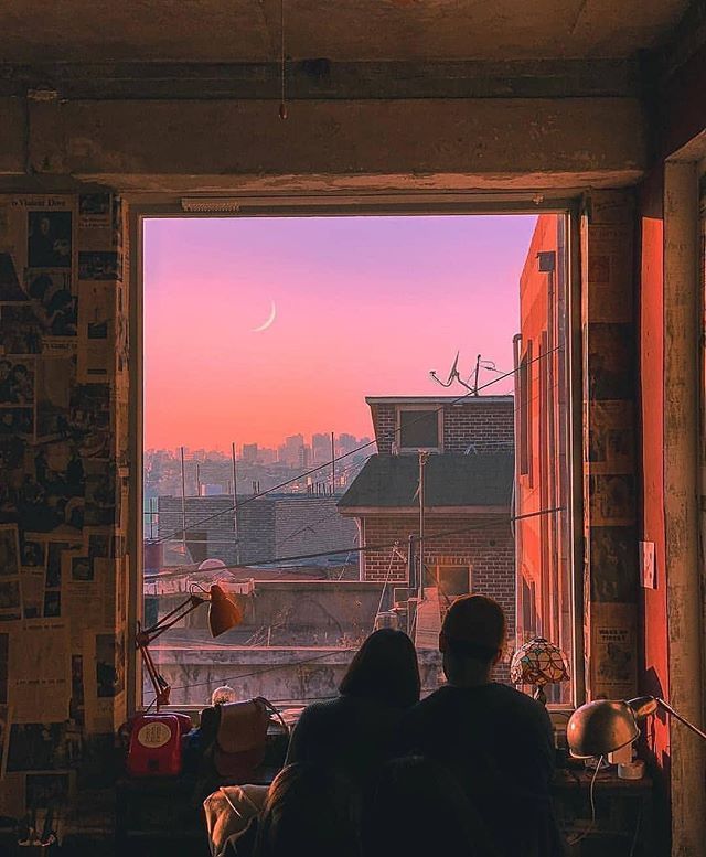 two people looking out an open window at the city below and pink sky in the distance