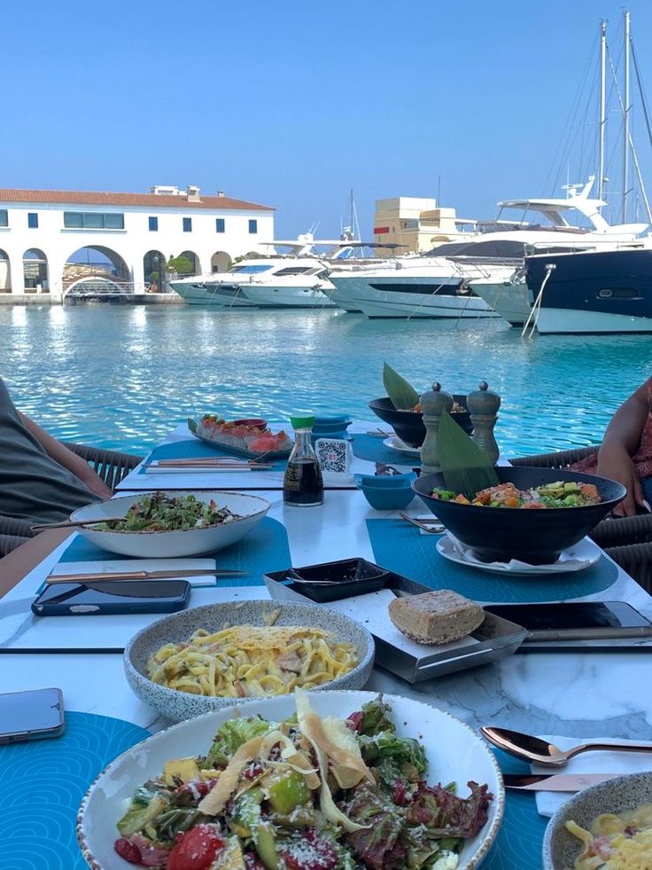 people sitting at a table with plates of food on it and boats in the water behind them