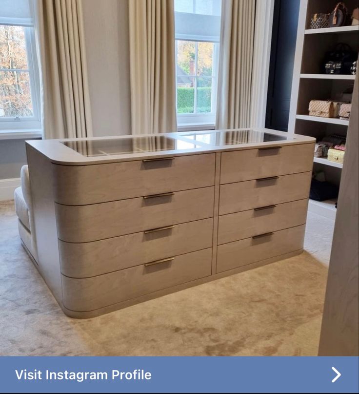 an empty dresser in front of two windows with curtains on the window sill behind it