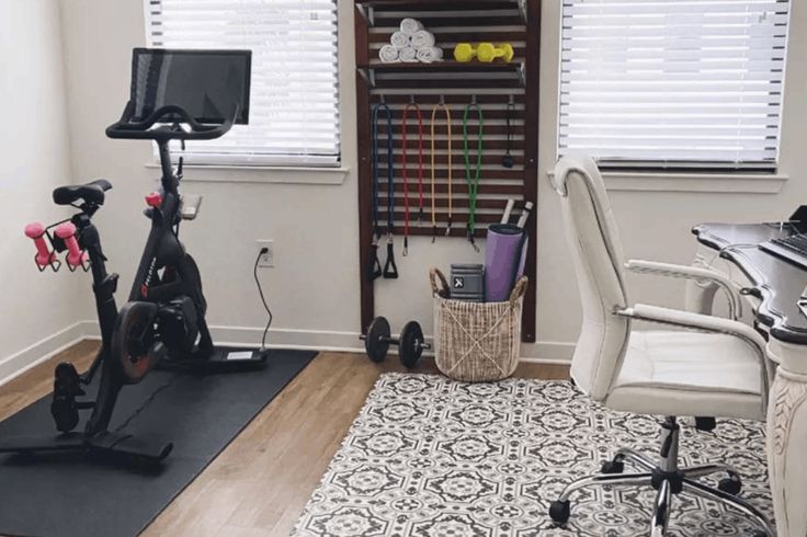 a home office with exercise bikes, desk and computer on top of a rug in front of the window