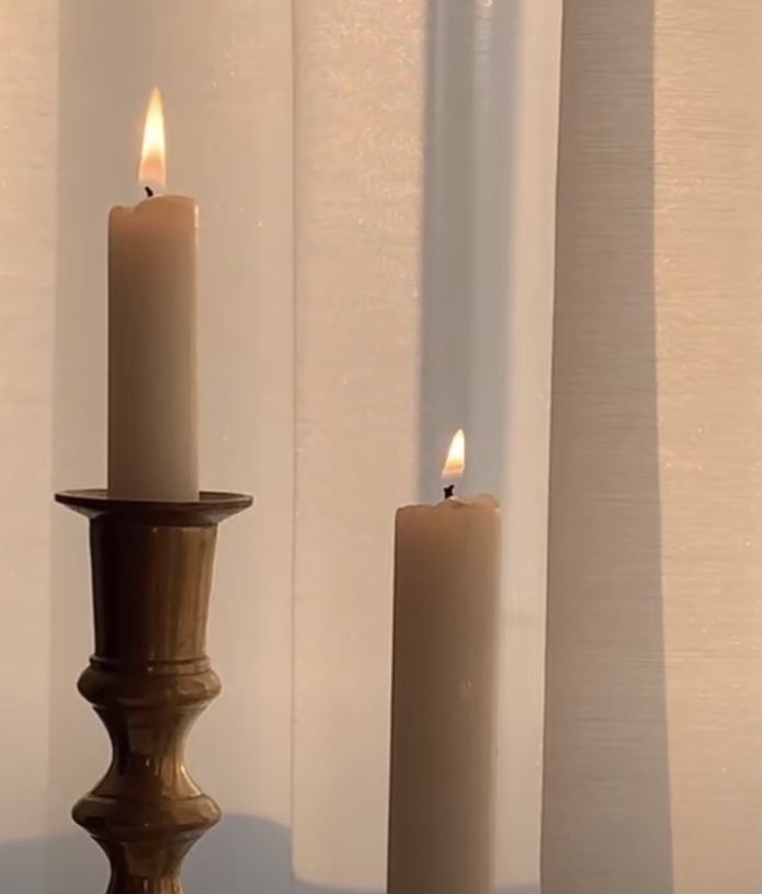 two lit candles sitting next to each other in front of a window with white curtains