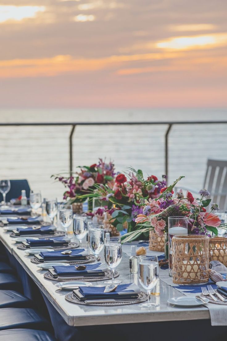 a long table set up with place settings and flowers on it, overlooking the ocean