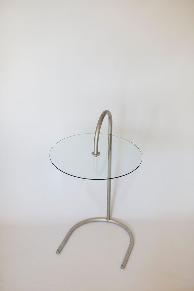 a round glass table with metal legs and a handle on the end, against a white wall