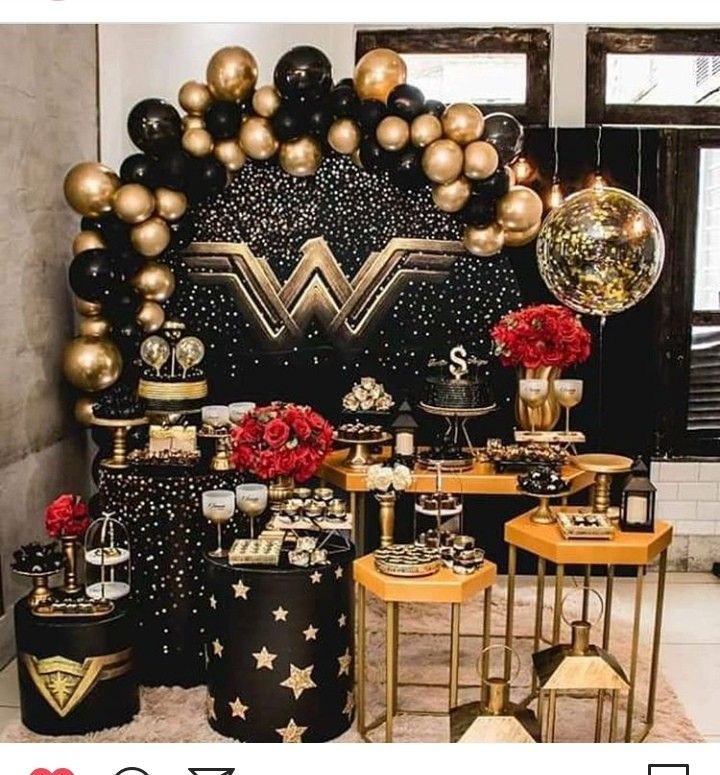 a table topped with lots of black and gold balloons next to a wonder woman backdrop