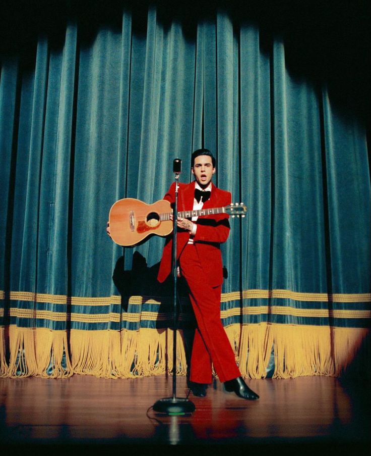 a man in a red suit playing an acoustic guitar on stage with curtains behind him