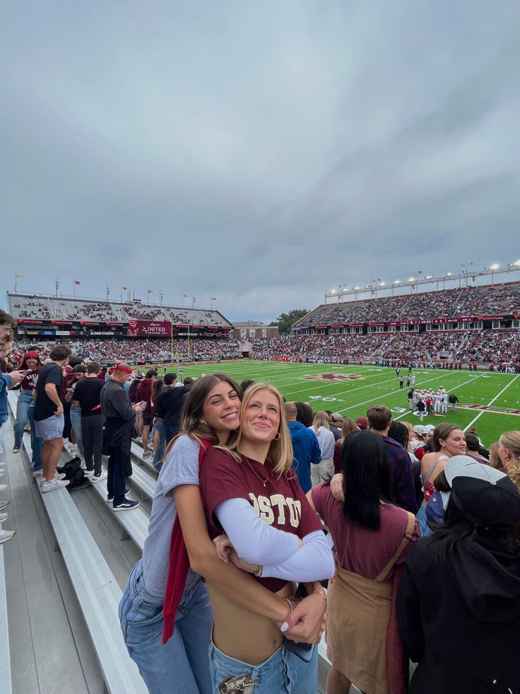 two women hugging each other in the stands at a football game with people watching from the bleachers