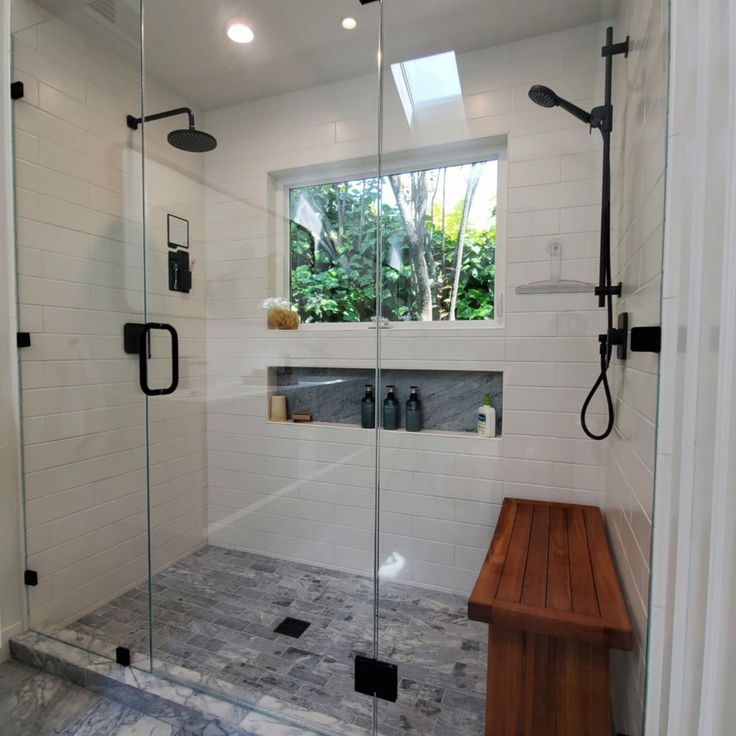 a walk in shower sitting next to a wooden bench under a large window on top of a white tiled wall