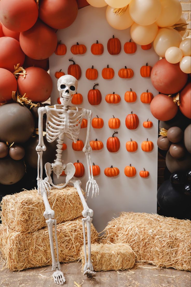 a skeleton sitting on hay bales in front of a pumpkin themed backdrop with balloons