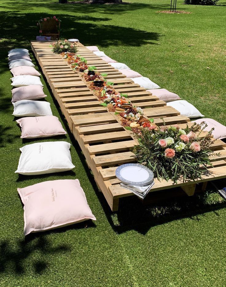 a long wooden table set up with pillows and flowers on the grass in front of it