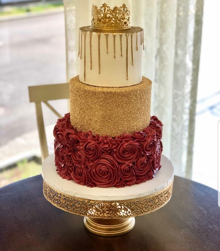 a three tiered cake with gold and red frosting on the top, sitting on a table in front of a window