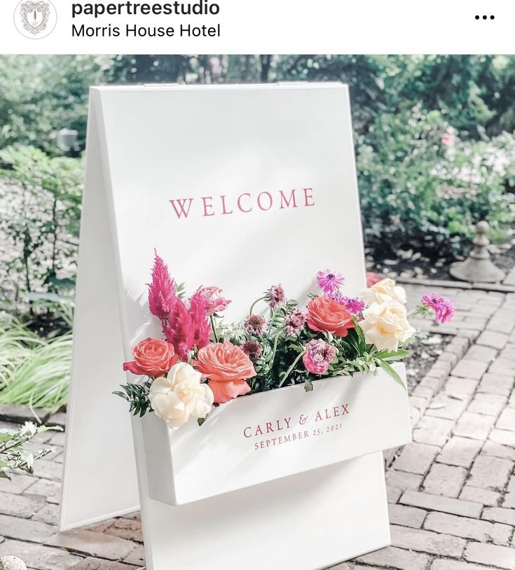 a welcome sign with flowers in it sitting on a brick walkway next to some bushes