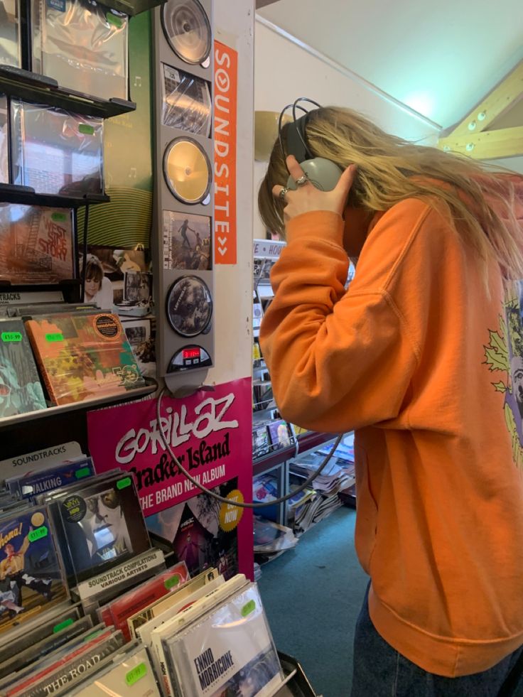 a woman in an orange sweatshirt is talking on her cell phone while she shops for cds