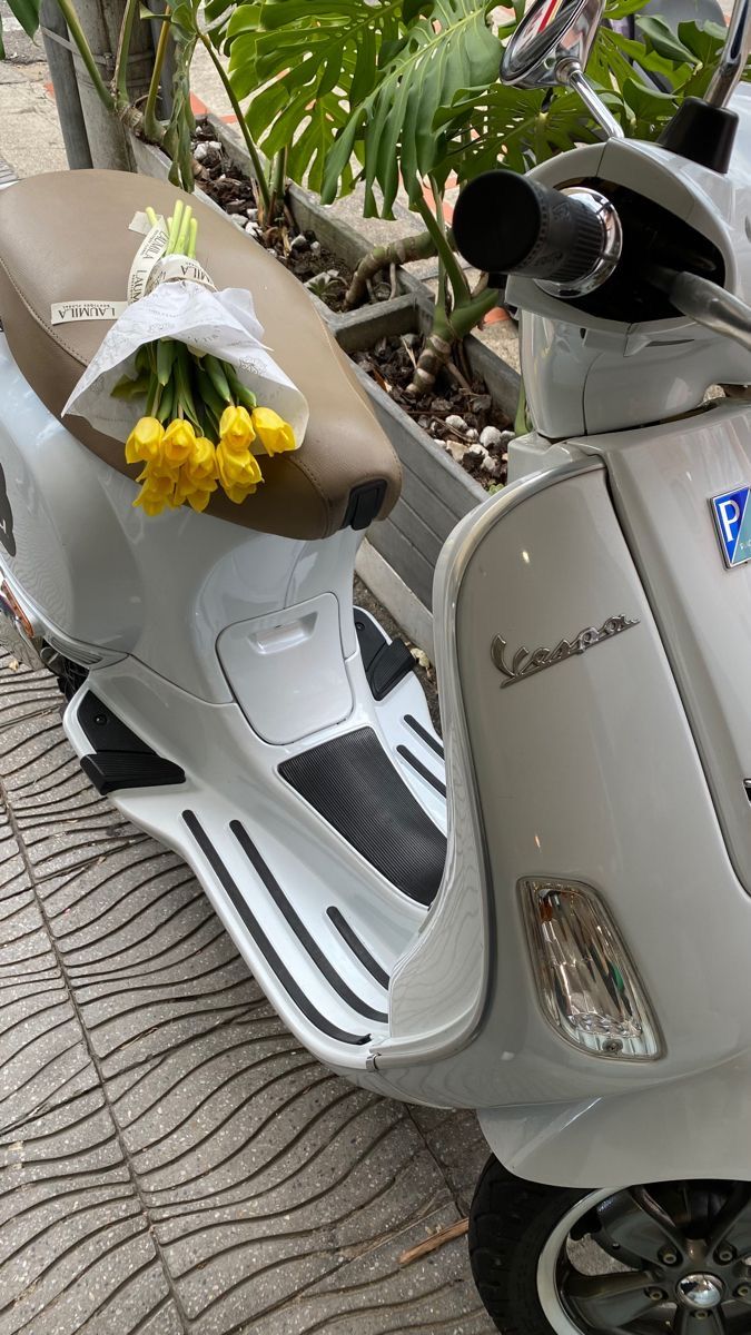 a scooter with flowers on the seat parked in front of a tree and plant