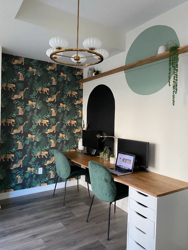 a desk with two green chairs and a laptop on it in front of a floral wallpaper