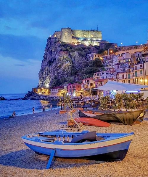 several boats on the beach with buildings in the background at night, and people walking around