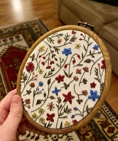 a hand holding a wooden frame with embroidered flowers on it in front of a couch