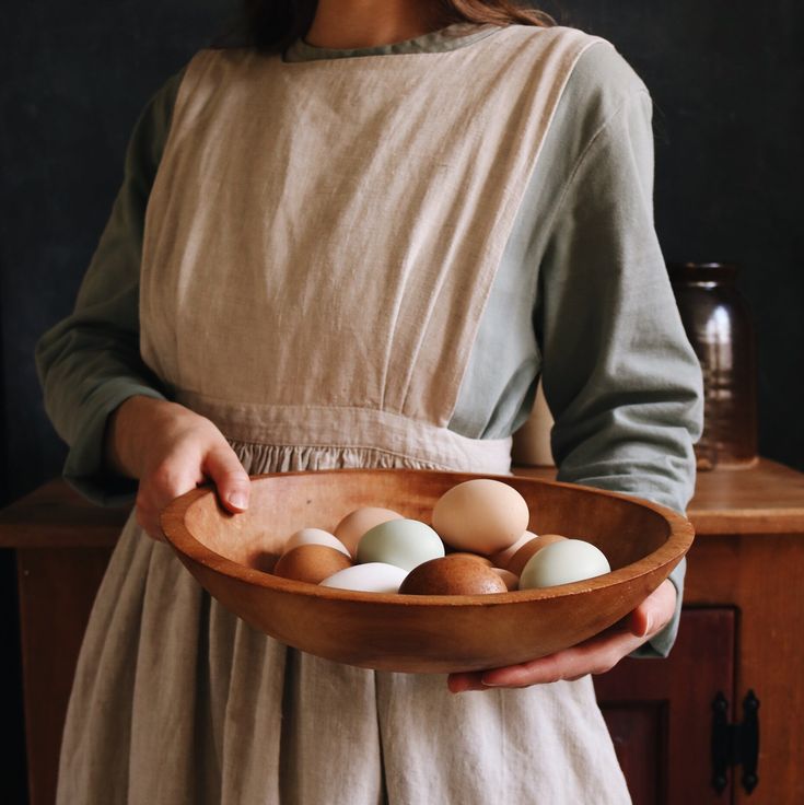 a woman in an apron holding a wooden bowl with eggs on it and one being held by her hand