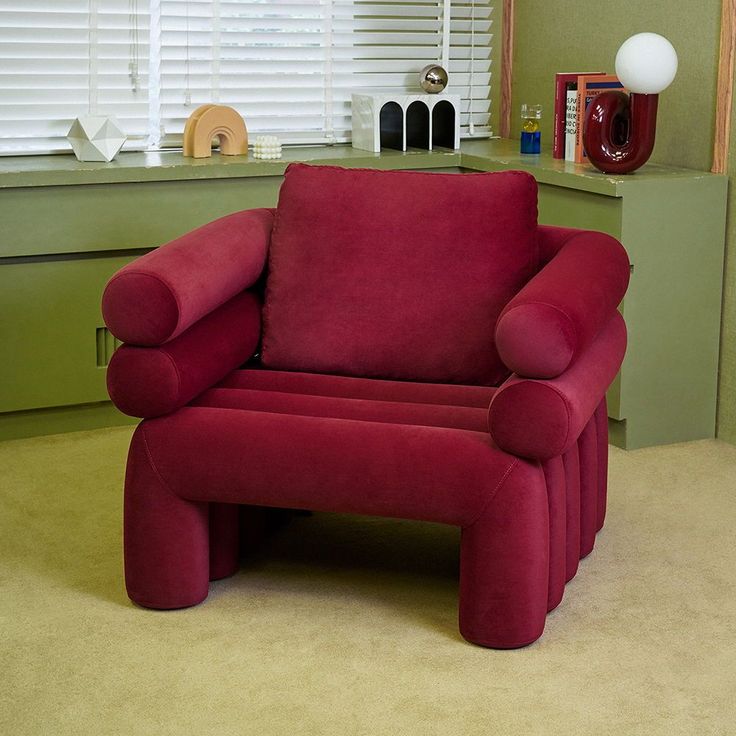 a red chair sitting in front of a window next to a lamp and bookshelf