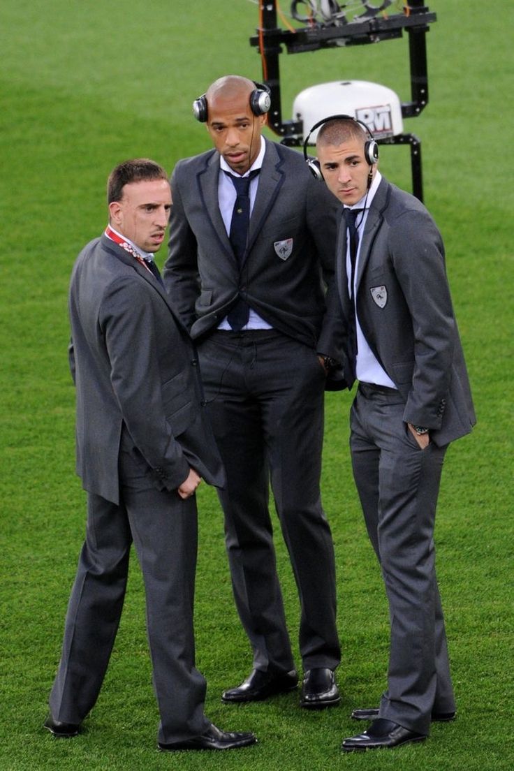 three men in suits standing on a soccer field next to a camera man wearing headphones