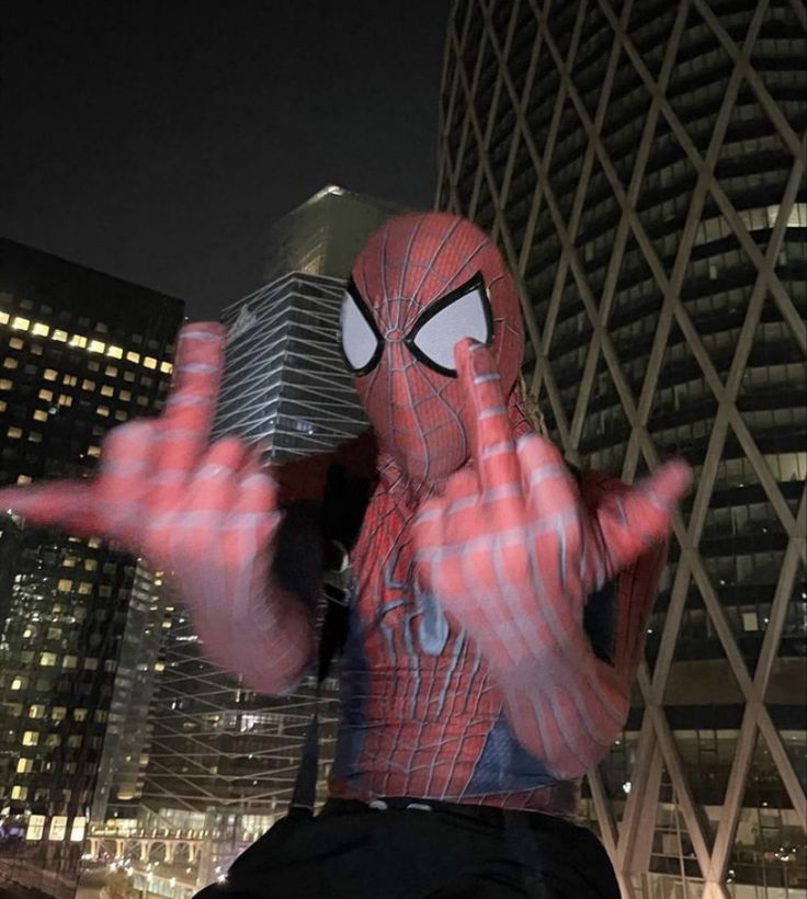 a spiderman statue in front of a skyscraper at night with his hands out to the side