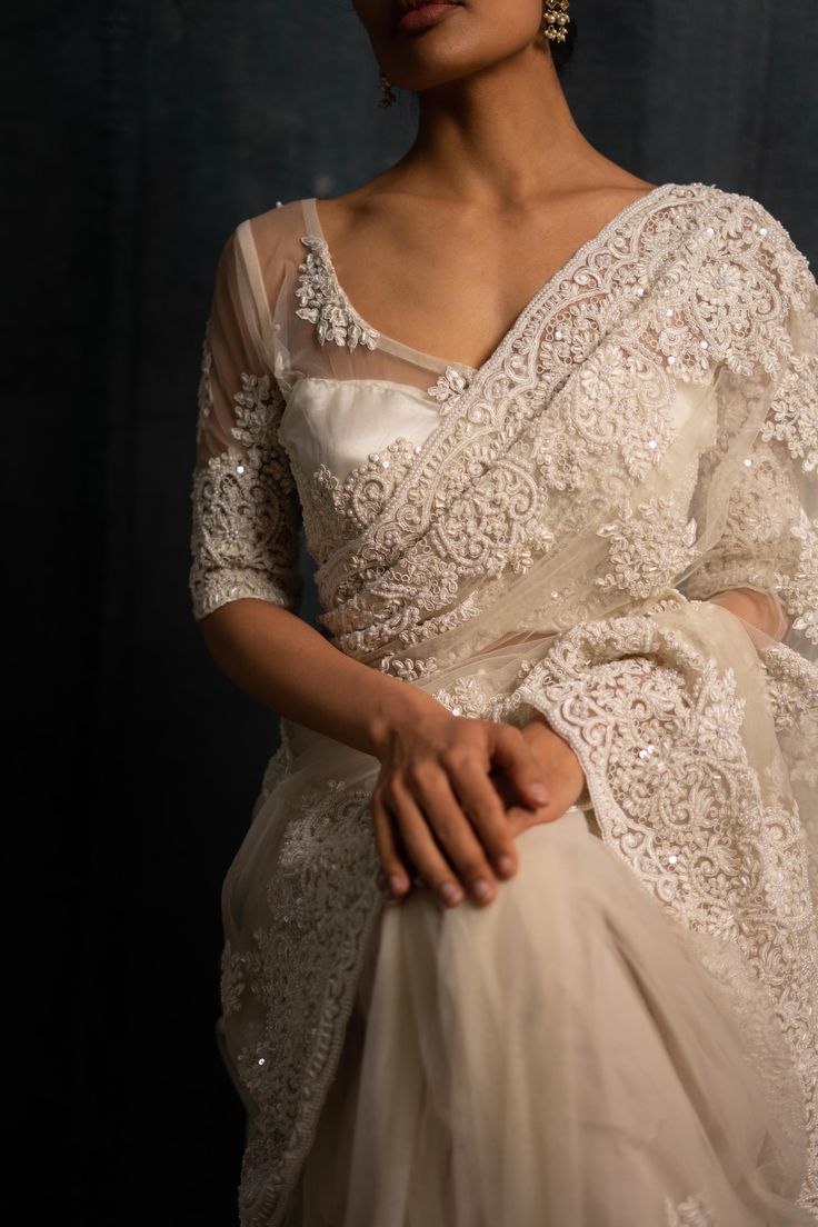 a woman wearing a white sari with intricate lace on the top and bottom, standing in front of a black background