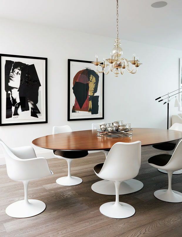 an oval dining table surrounded by white chairs and art on the wall behind it, with chandelier hanging above