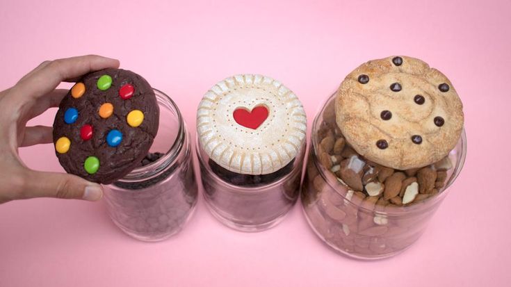 three jars filled with cookies and candy on top of a pink background, one holding a cookie in the other's hand