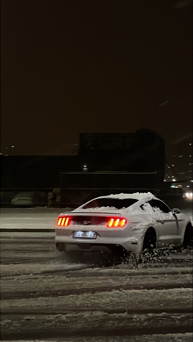 a white car driving down a snow covered road at night with buildings in the background