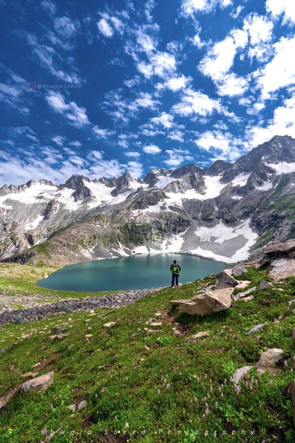 a man standing on top of a lush green hillside next to a lake in the mountains