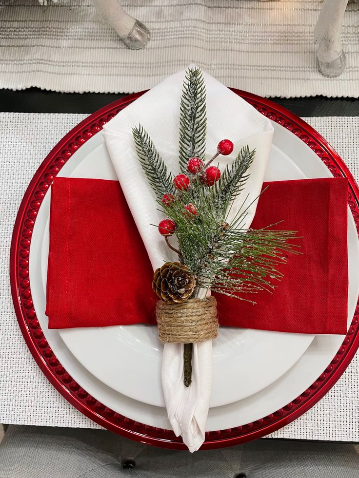 a red and white place setting with pine cones, greenery and berries on the napkins