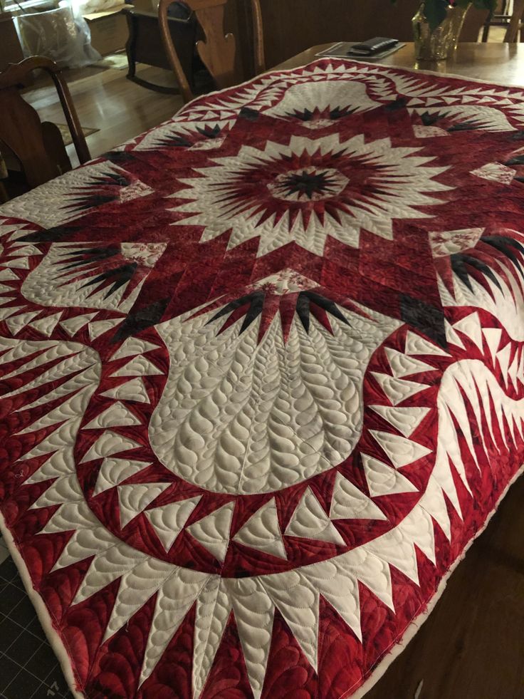 a large red and white quilt on top of a table with chairs in the background