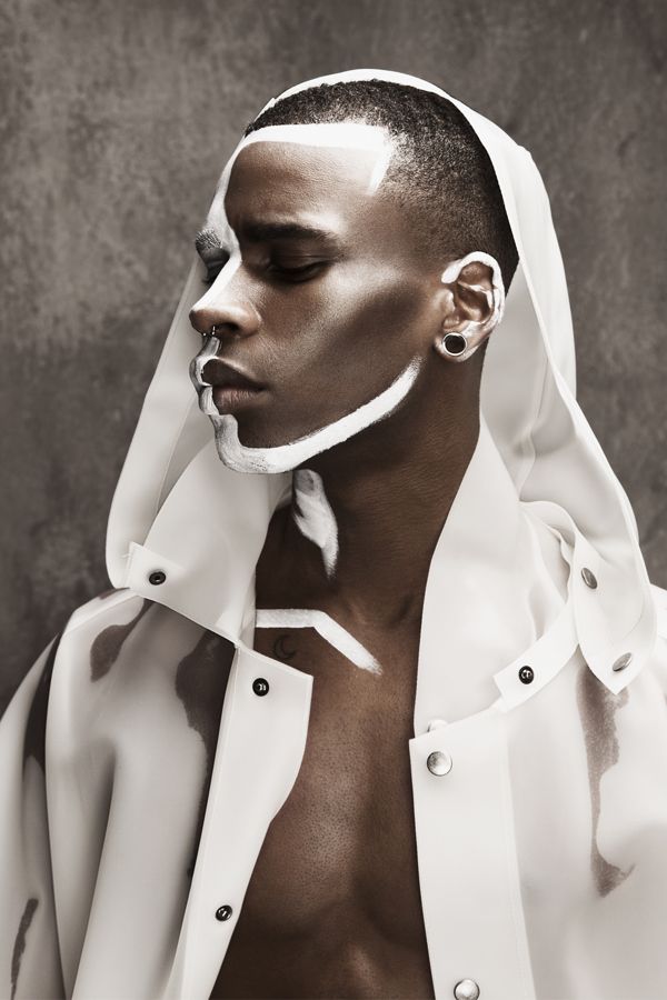 a man with no shirt on wearing a white jacket and silver earrings, standing in front of a gray background