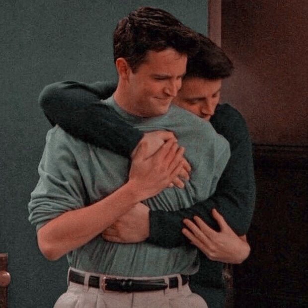 two men hug each other while standing in front of a green wall, one holding his arm around the other