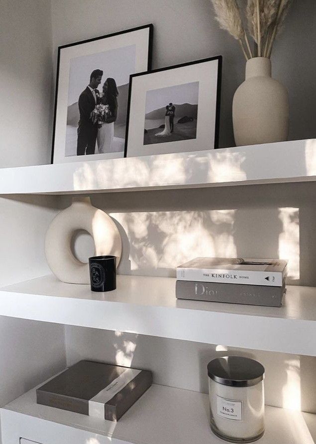 some books and pictures on a shelf in a room with white shelves, two vases and one candle