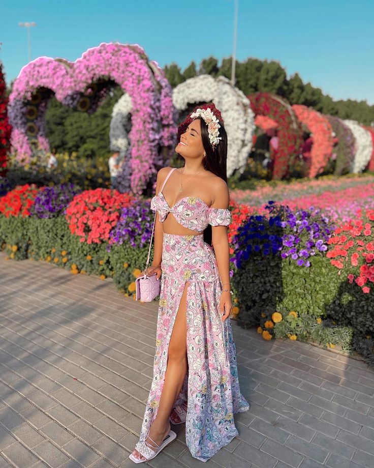 a woman standing in front of flowers wearing a floral dress and holding a pink purse